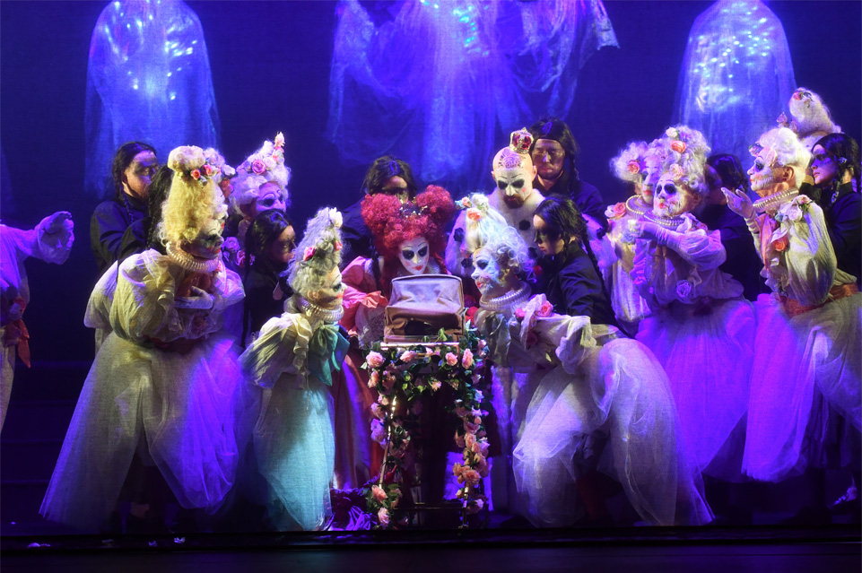 A group of performers, dressed in fantastical costume, surrounding a crib, with well-lit fairies in the background, performing for an audience.